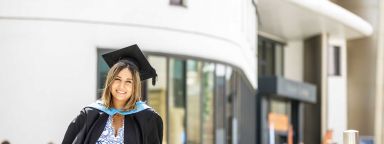 Smiling graduate wearing a cap and gown outside a university building.