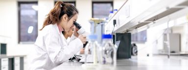 Woman in a white lab coat looking through a microscope in a laboratory.